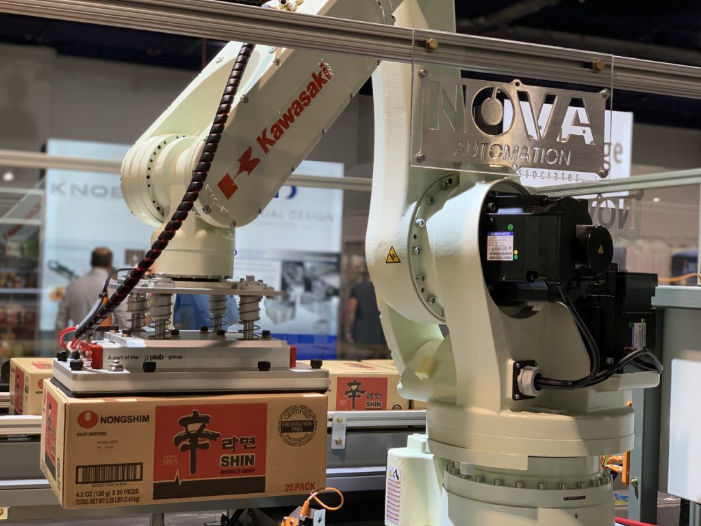 A Kawasaki robotic arm is lifting a box of Nongshim Shin Ramyun noodles, at an INOVA AUTOMATION exhibit. There are blurred booth displays in the background.