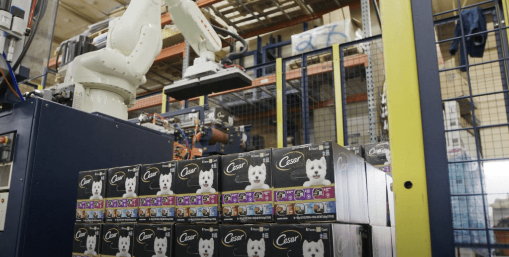 A robotic arm is handling boxes of Cesar dog food in a manufacturing facility with industrial shelving and equipment in the background.