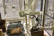 A robotic arm is positioned above a table, seemingly interacting with blocks or components, in a mesh-enclosed industrial space with wiring and metallic structures.
