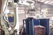 A large industrial robot arm is positioned near stacks of bagged products inside a warehouse with metal structures and machinery.
