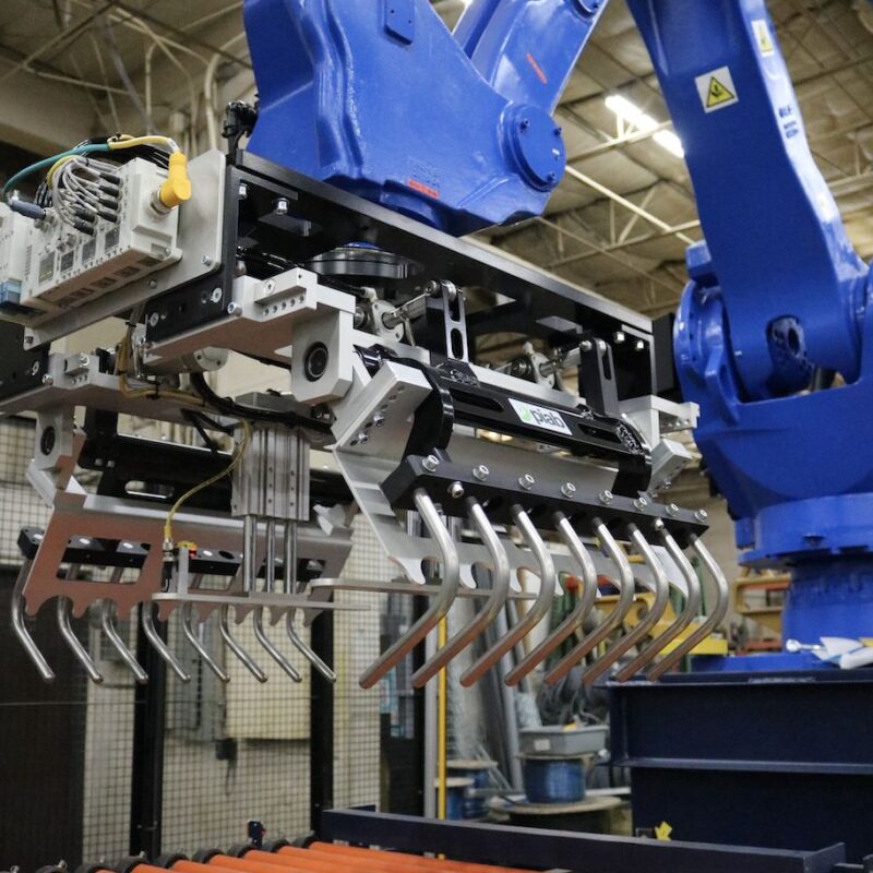 A blue industrial robot with a multi-clawed attachment is positioned above a conveyor belt with orange rollers in a factory setting.
