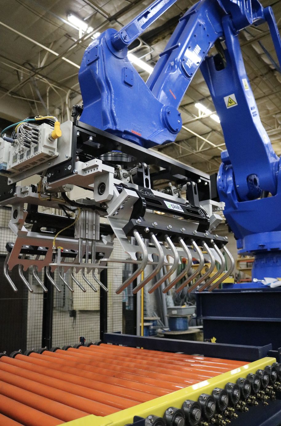 A blue industrial robot with a multi-clawed attachment is positioned above a conveyor belt with orange rollers in a factory setting.